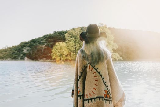 Boho style woman wearing in poncho and hat walking on lake, rear view.