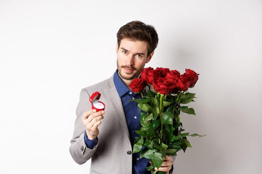 Handsome man in suit, showing engagement ring and looking romantic at camera, standing with red roses over white background. Valentines day and love concept.
