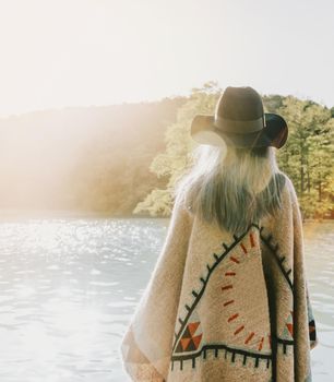 Boho style girl wearing in poncho and hat walking on lake, rear view.