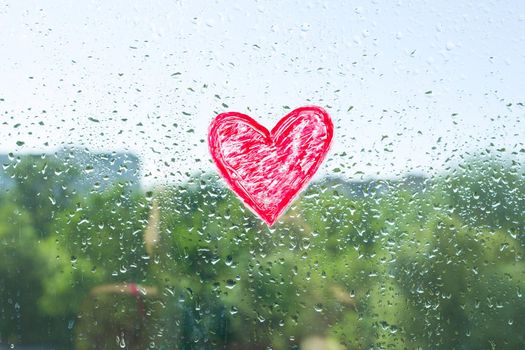 Red heart painted with lipstick on the window with water drops. Background blue sunny sky, drops shine in the sun