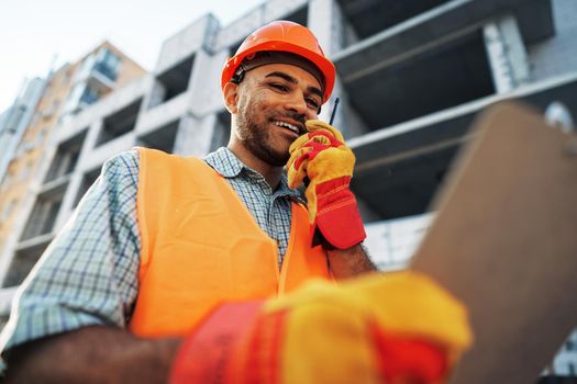 Young construction worker in uniform using walkie talkie on site, close up portrait