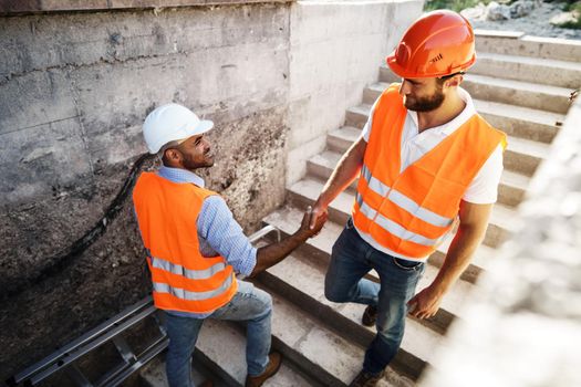 Two men engineers in workwear shaking hands against construction site, close up