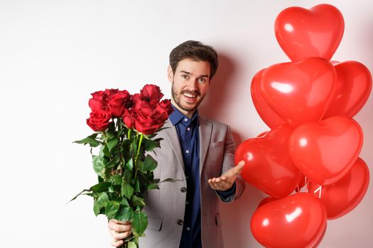 Romantic guy in suit saying happy Valentines day, giving red roses to you and pointing hand, standing with heart balloon gift over white background.