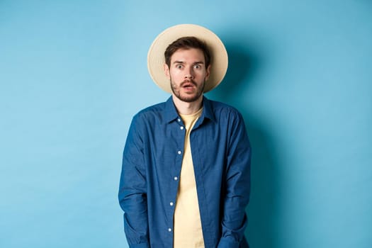 Young man tourist in summer hat drop jaw, staring at something shocking, standing on blue background.