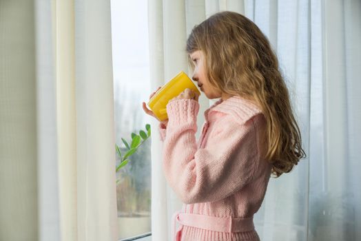 Girl child 7 years old blonde with long wavy hair in a warm knitted sweater holds a cup of tea and looks in the window