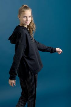 Pretty teenage girl in black stylish clothes. Portrait of adorable girl with ponytail dressed sweatshirt and plaid trousers standing against blue background in studio staring at camera