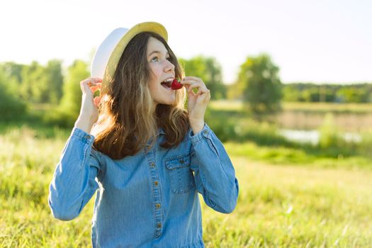 Attractive teen girl eating strawberry. Nature background, rural landscape, green meadow, country style.