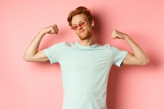 Confident young man with red hair, wearing summer sunglasses and t-shirt, showing strong and fit body muscles, flex biceps and staring cool at camera, pink background. Workout and gym concept