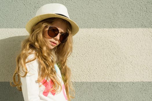 Outdoor portrait little blonde girl with long curly hair, sunglasses in straw hat. Gray textured wall background, copy space.