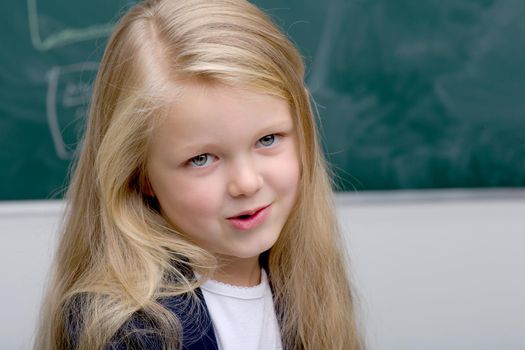 Close up portrait of beautiful girl. Adorable blonde long haired girl posing on background of blackboard in classroom. Cute smiling elementary school student girl. Back to school, education concept