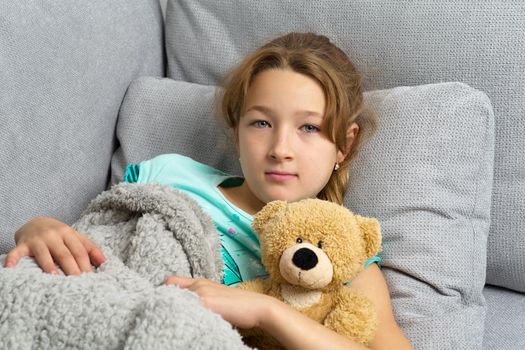 Beautiful blonde girl lying on soft comfortable sofa with fluffy teddy bear. Portrait of relaxed teen girl lying covered with plaid. Child resting on couch hugging favorite stuffed toy