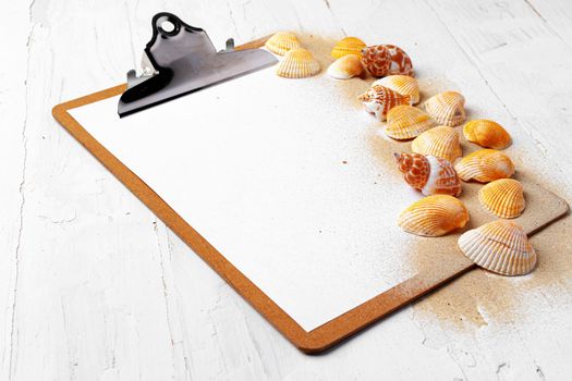 Clipboard with sea shells on white wooden table close up, copy space.