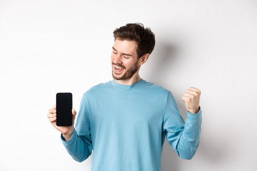 E-commerce and shopping concept. Lucky man winning online, showing empty smartphone screen and triumphing, saying yes with fist pump, standing over white background.