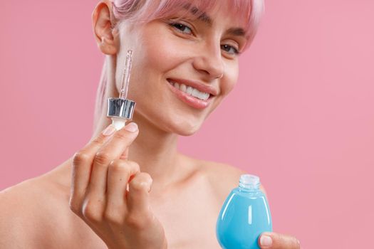 Portrait of young woman with pink hair holding dropper and a bottle of serum or hyaluronic acid, posing isolated over pink background. Beauty, cosmetics and skincare concept