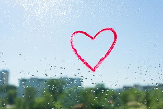 Red heart painted with lipstick on the window with water drops. Background blue sunny sky, drops shine in the sun