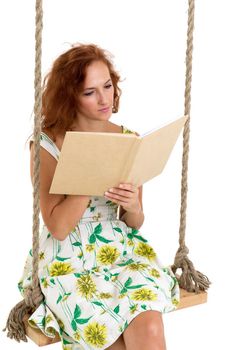 Happy young woman reading book on swing. Smiling woman wearing summer dress sitting on rope swing against white background. Portrait of beautiful girl posing in studio