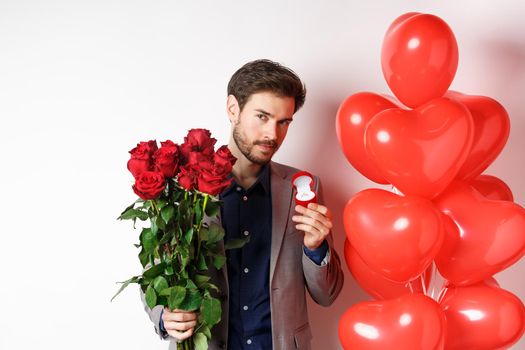 Handsome boyfriend in suit making proposal on lovers day, holding engagement ring and red roses, prepare surprise flowers and heart balloons for girlfriend on Valentines, white background.