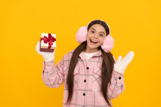 teen girl in mittens hold present on yellow background. xmas holiday gift. child wearing warm clothes. boxing day. happy new year. merry christmas. happy amazed kid in winter earmuffs with box.