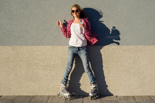 Portrait of young cool smiling girl shod in rollerblades, holding a water bottle and showing a thumbs up. Thumb up health and sport