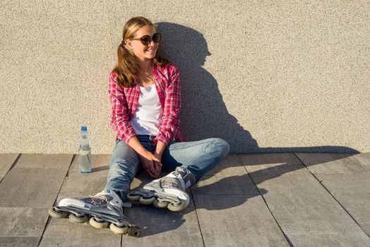 Young smiling cool girl shod in rollerblades, sits on the sidewalk and holding a water bottle. Sports, health and active leisure.