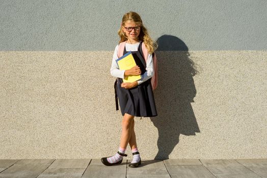 An elementary school student with notebooks in his hand. A little schoolgirl with a backpack near the building in the open air.