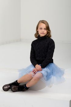 A beautiful girl in a black jumper and a blue fluffy skirt sits on a white staircase. Pretty blonde woman with stylish hairstyle in fashionable clothes posing in studio