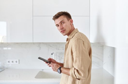 Hadsome smiling man is chilling on the minimalistic kitchen with smartphone