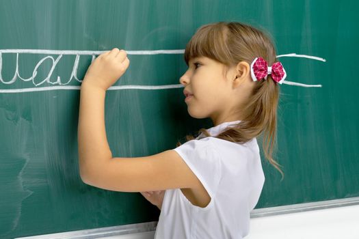 Cute schoolgirl writing on blackboard. Portrait of elementary school student girl in white blouse standing next to chalkboard in classroom. School and education concept