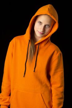 Shot of beautiful stylish teenage girl. Portrait of attractive teenager wearing warm orange hoodie putting the hood on. Beautiful brown eyed girl posing against black background