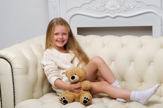 Lovely blonde girl sitting on comfortable sofa hugging teddy bear. Portrait of cute long haired girl wearing warm knitted dress. Adorable happy child resting on couch with favorite stuffed toy