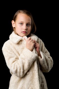 Close up portrait of stylish preteen girl. Beautiful fashionable blonde girl wearing beige faux fur coat posing against black background. Trendy child dressed fashion outfit