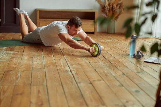 Sporty guy is using abs wheel in living room while watching online workout on notebook