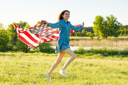 Girl teenager with American flag running through the summer green meadow at sunset. Nature background, rural landscape.