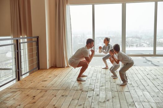 Athletic man is showing his wife and son with daughter how to exercise before window indoors