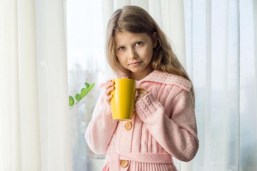 Girl child 7 years old blonde with long wavy hair in a warm knitted sweater holds a cup of tea and looks in the window