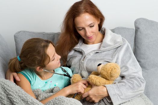 Mom looking lovingly at her daughter. Mother and child playing doctor and patient sitting together on couch at home. Cheerful woman using stethoscope to listen teddy bear toy heart