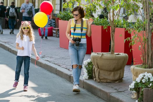 Urban portrait of mother and daughter. Girl with balloons is walking along the city street.