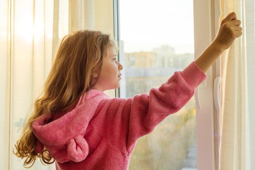 Good morning. Girl child in pajamas opens curtains and looks out the window