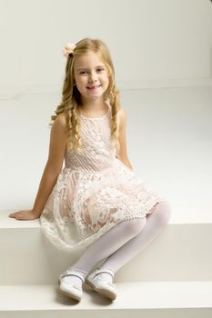 Lovely girl sitting on white staircase. Portrait of beautiful blonde girl wearing nice dress, tights and shoes posing in studio. Lovely smiling seven years old kid sitting on white stairs