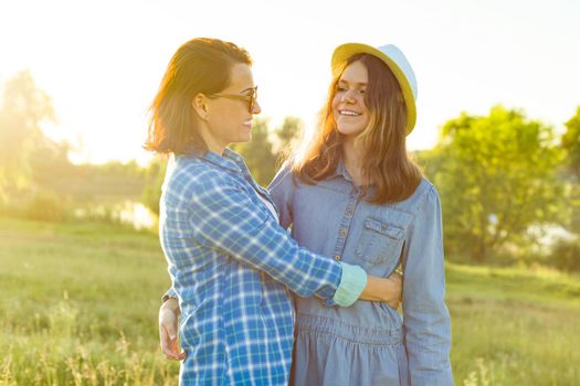 Parent and teenager, mother and 14 year old daughter embrace smiling in nature. Background sunset, rustic landscapes, green meadow