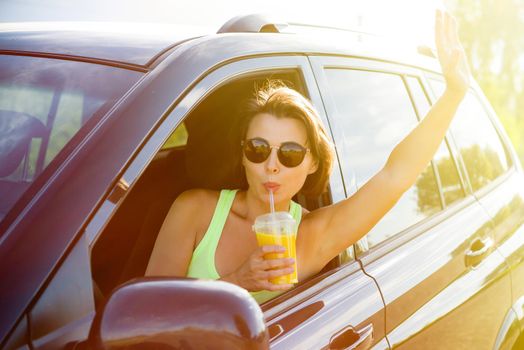 A happy woman driver stops to relax, enjoy the journey, drink orange juice and wave your hand