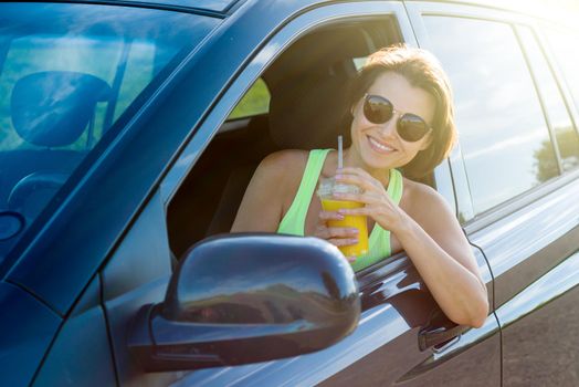 Beautiful woman is drinking juice, looking at camera and smiling while sitting in her car