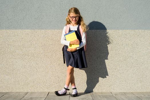 A pupil of elementary school with notebooks in his hand. Girl with a backpack near the building outdoors. Start of classes.