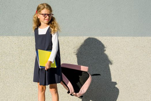 A schoolgirl of primary school with notebooks in his hand. Girl with a backpack near the building outdoors. Back to school!