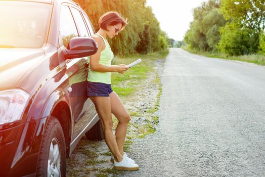 Cute female driver is traveling. Standing beside his car on a country road and watching a road map.