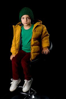 Cute boy in winter jacket and knitted hat. Stylish cheerful boy sitting on chair posing at camera in studio. Child wearing fashionable winter outerwear isolated on black background