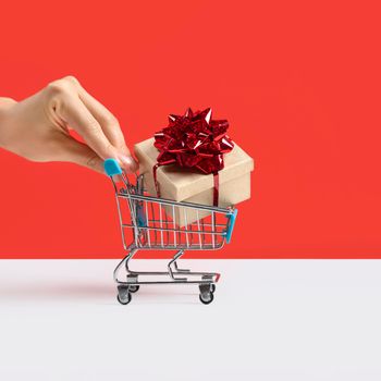 Hand pushing Toy shopping cart or trolley with large gift box. A gift for a holiday, anniversary, or birthday. Trolley cart with a gift box Isolated over red background