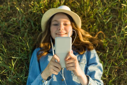 Teenage girl in hat with headphones lies on the green grass and looks into the phone. Focus on smartphone, top view