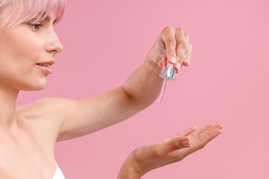 Close up portrait of happy young woman with pink hair and perfect skin holding dropper with serum or hyaluronic acid to apply it on skin, posing isolated over pink background. Side view