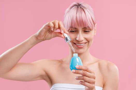 Portrait of happy young woman with pink hair and bare shoulders smiling and holding bottle of serum, posing isolated over pink background. Beauty, cosmetics and skincare concept